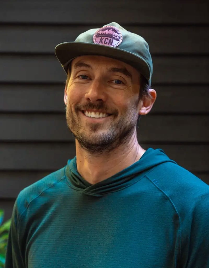 A man in a green hoodie and cap smiles in front of a dark wooden wall.