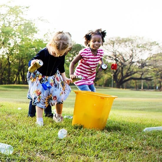 Two young girls pick up plastic bottles and cans near a large yellow bin on a grassy field, participating in an outdoor cleanup activity.