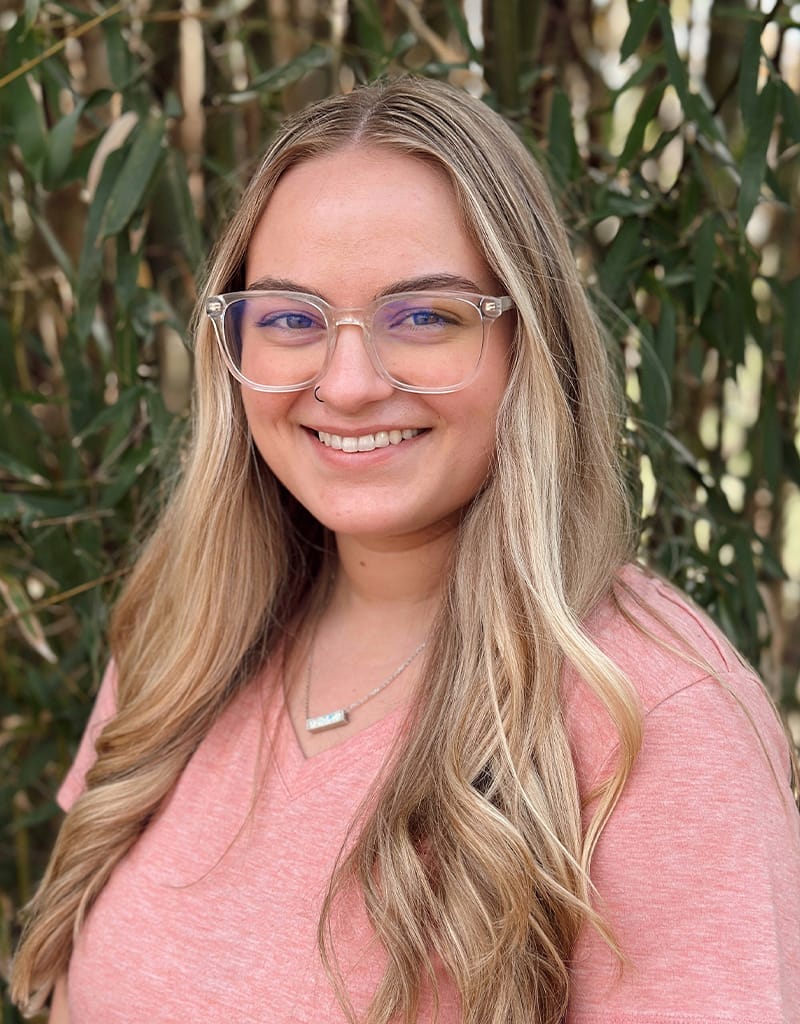 Lexie has long blonde hair and glasses and is wearing a pink shirt.