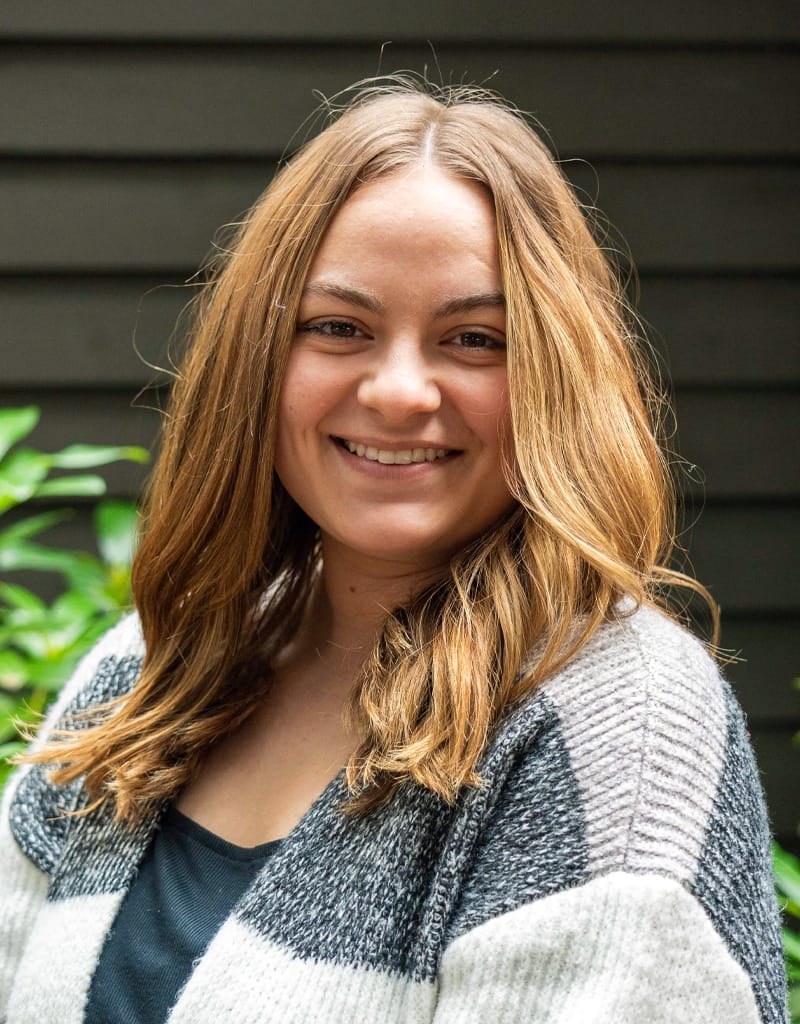 A woman with long brown hair smiles, wearing a gray and black sweater, standing in front of a dark wall with some greenery visible.