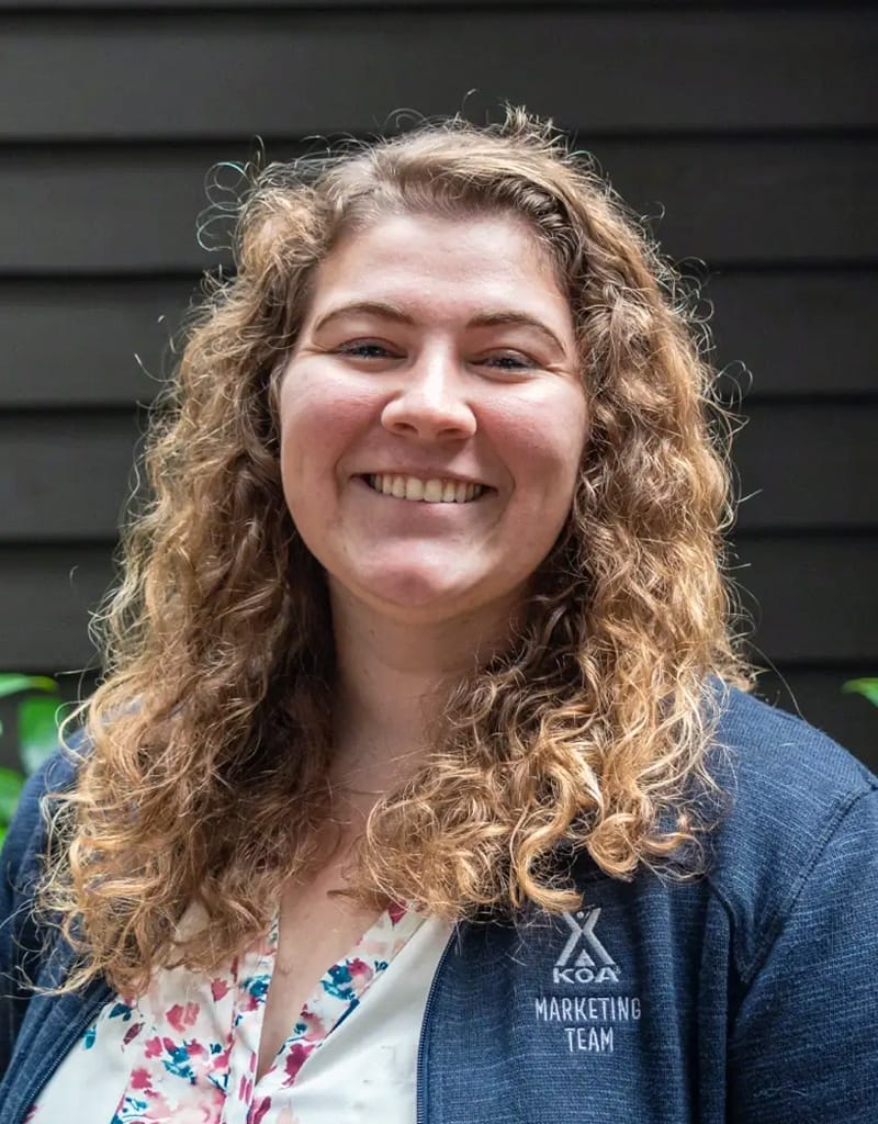 Woman with curly hair smiling, wearing a jacket with "Koa Marketing Team" logo, stands in front of a dark wood wall.