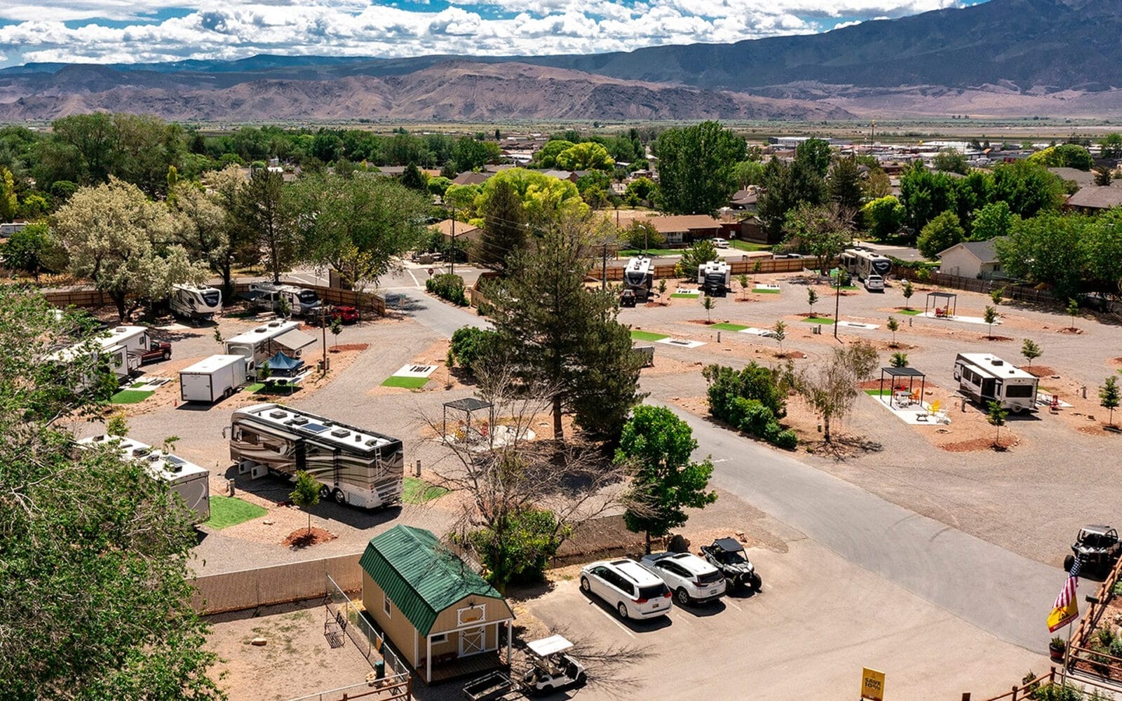 Aerial view of Richfield KOA campground with several RVs, scattered trees, and pathways. Surrounding mountains and a partly cloudy sky are in the background.