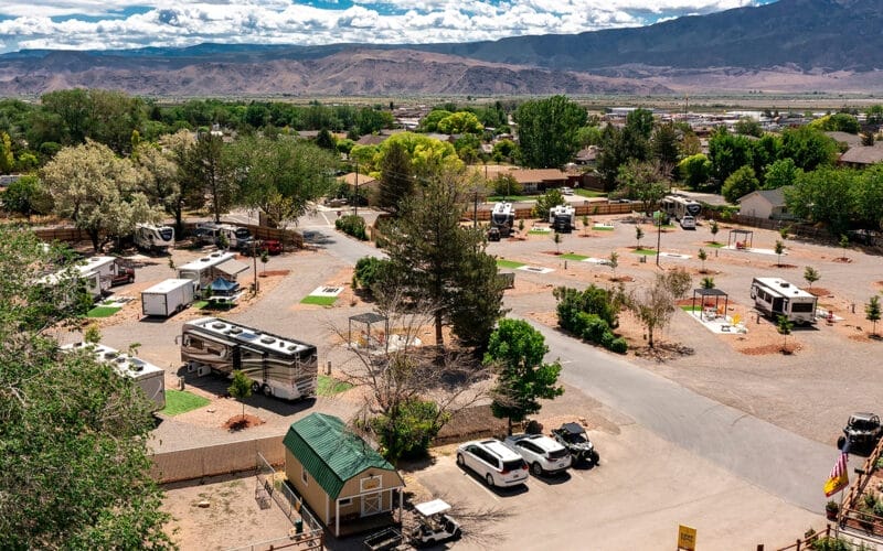 Aerial view of Richfield KOA campground with several RVs, scattered trees, and pathways. Surrounding mountains and a partly cloudy sky are in the background.