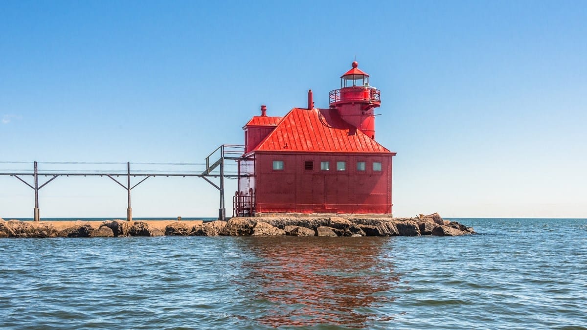 A bright red lighthouse with an attached walkway sits on a rocky pier extending into a calm body of water under a clear blue sky at Sturgeon Bay.