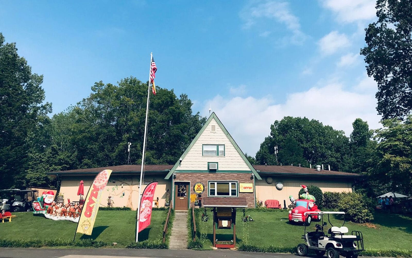 Sweetwater KOA building with flags, cartoon car decoration, and golf cart on a grassy yard with trees in the background.