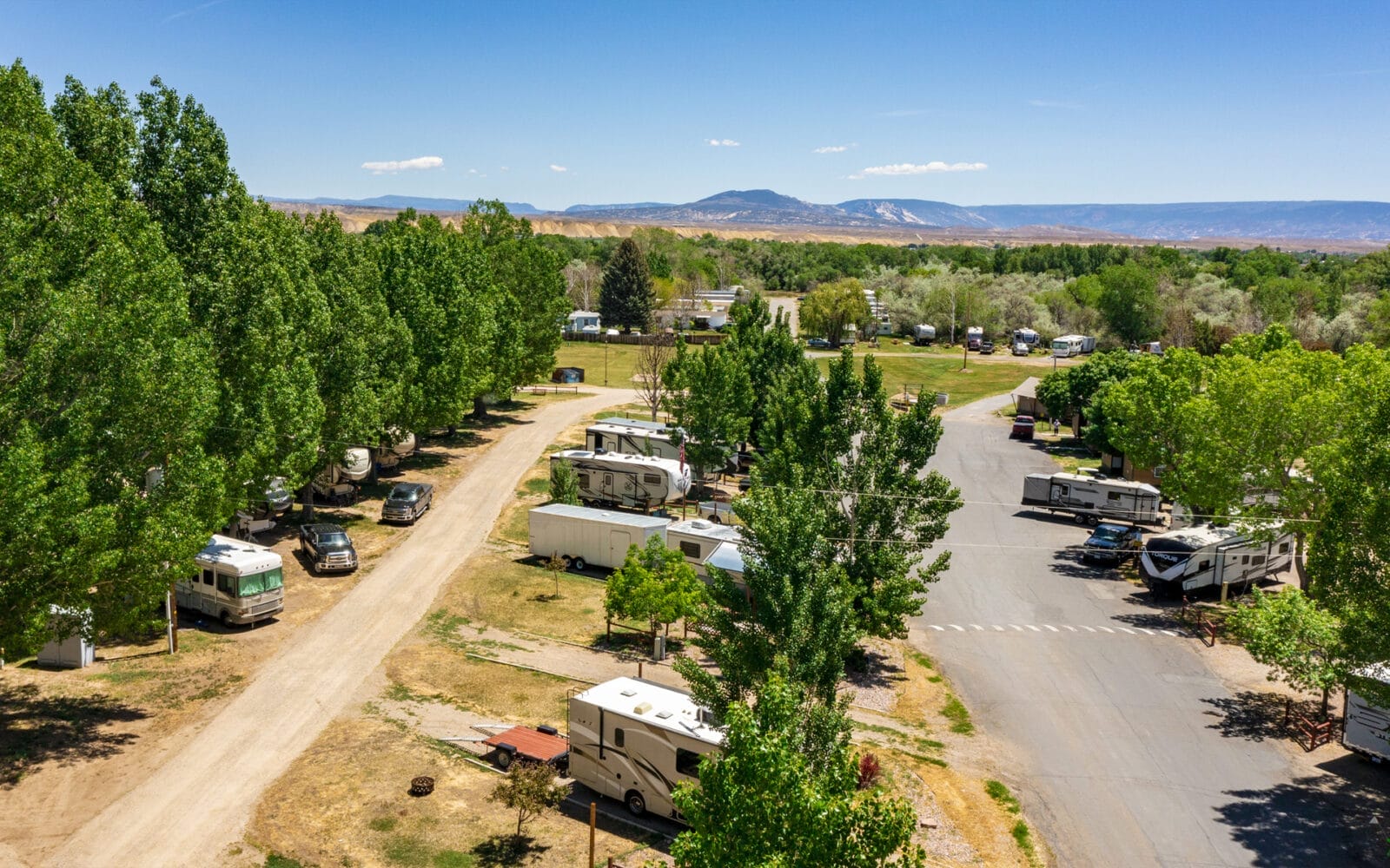 Aerial view of a Vernal / Dinosaurland KOA campground with RVs and trailers among trees, a dirt road to the left, paved road to the right, and mountains in the background under a clear blue sky.